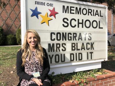 Memorial Elementary School kindergarten teacher Samantha Black sits next to a Memorial School marquee, saluting her selection as the 2019 recipient of the Cole Chevrolet Four Seasons Teacher of the Year award.